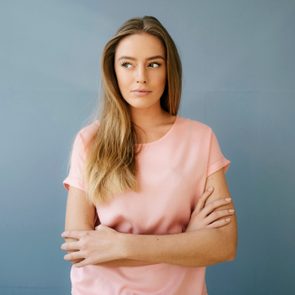 Woman stands with arms crossed, gazing sideways, wearing a pink shirt against a plain blue background.