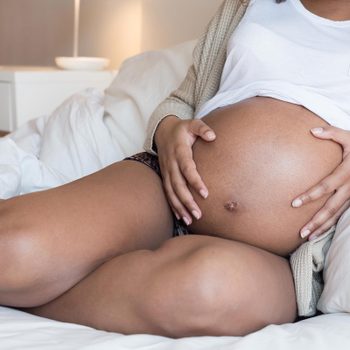 Pregnant woman rests hands on her belly while sitting on a bed with white sheets in a softly lit room.