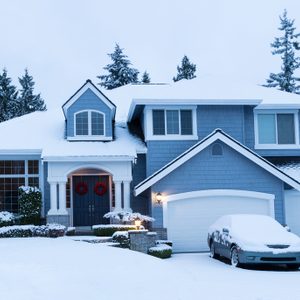 House stands snow-covered, displaying holiday wreaths; car parked in driveway similarly blanketed. Snow-dusted trees frame the suburban scene.