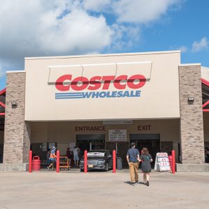 Costco entrance features people walking inside a brick and concrete facade under a cloudy sky.