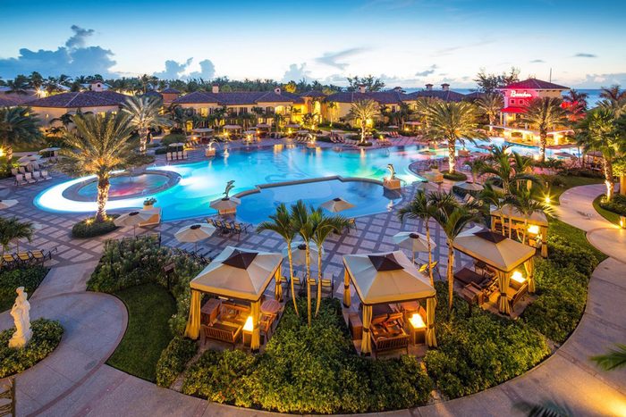 A large, illuminated pool surrounded by cabanas and palm trees at a resort during twilight, creating a tranquil, luxurious ambiance.