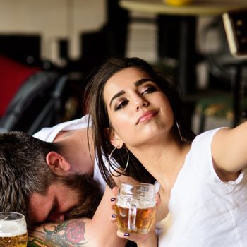 Woman takes a selfie, holding a beer mug, while man sleeps on table, indoors at a bar or restaurant setting.