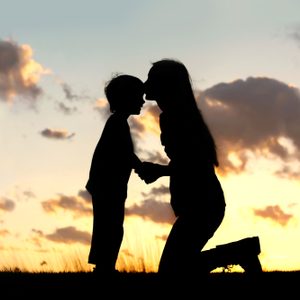 Silhouetted figures embrace at sunset, one kneeling, one standing, against a backdrop of vivid clouds and warm, glowing sky.