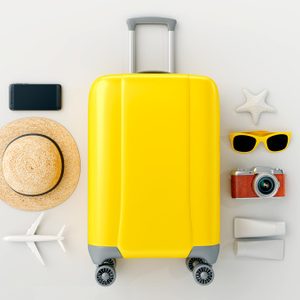 Yellow suitcase surrounded by camera, sunglasses, hat, smartphone, starfish, airplane model, sunscreen on a light background.