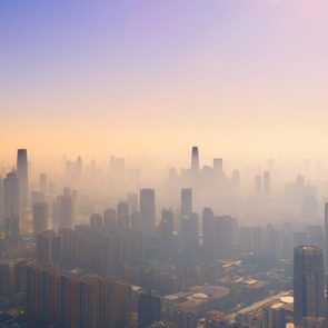 Skyscrapers rise, silhouettes shrouded in mist, against an orange-pink gradient sky in a sprawling urban skyline.