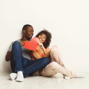 A couple sits on the floor, smiling and holding a red paper heart against a plain white wall.