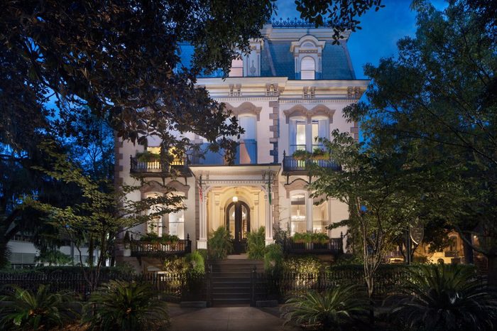 A Victorian-style house stands illuminated at night, surrounded by lush trees and a wrought-iron fence, with glowing windows highlighting intricate architectural details.