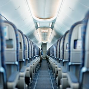 Rows of empty airplane seats stretch along a narrow aisle, under overhead bins and illuminated by soft ceiling lights. An 