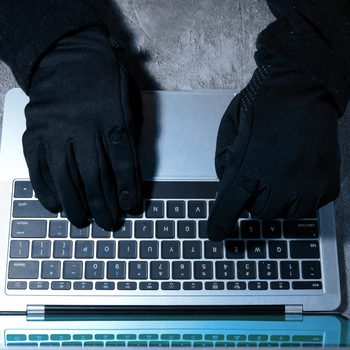 Gloved hands typing on a laptop against a textured gray surface, suggesting an anonymous or secretive activity.