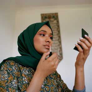 Person applies lip gloss while holding a small mirror in a softly lit room with decorative wall hanging.