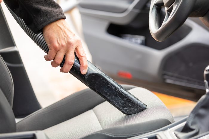 Hand vacuuming car seat with a nozzle, inside a vehicle's interior, near the steering wheel and dashboard area.
