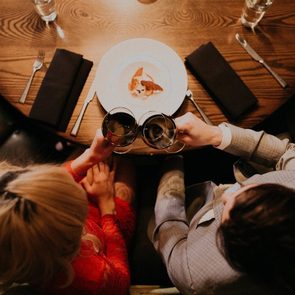 Two glasses clink above a plate at a restaurant table, surrounded by cutlery, napkins, and water glasses.