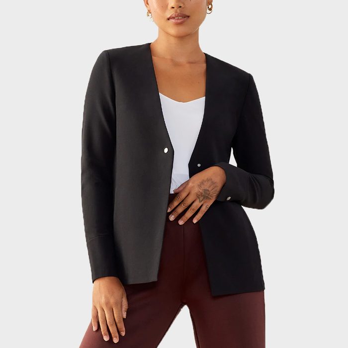 A person stands wearing a black blazer over a white top and burgundy pants, in a neutral setting.