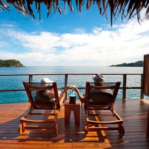 Two people relax on lounge chairs, clinking glasses, overlooking a serene ocean from a wooden deck under a thatched roof.