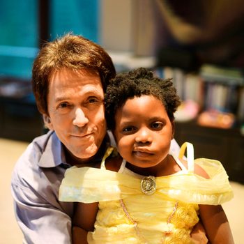 A man hugs a young girl in a yellow dress, inside a room with blurry bookshelves in the background.