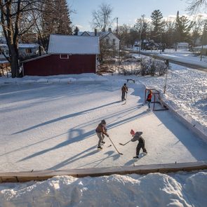 People play ice hockey on a backyard rink, surrounded by snow and trees, with houses and clear skies in the background.