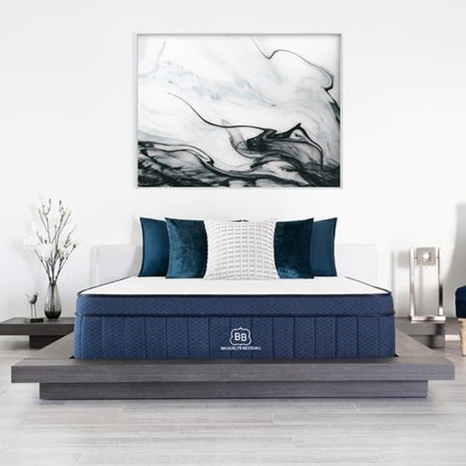 Bed with blue bedding and pillows sits in a modern, minimalist bedroom with abstract wall art and small decorative plants on side tables.