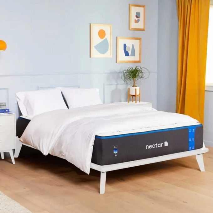 Bed is neatly made with white bedding, inside a bright room featuring abstract wall art, a potted plant on a nightstand, and yellow curtains.