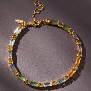 Bracelet, featuring colorful gems in gold settings, rests on a textured dark surface, casting soft shadows.