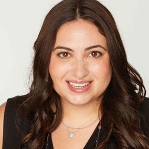 A person smiles directly at the camera. They have long, wavy hair and are wearing a necklace. The background is plain white.