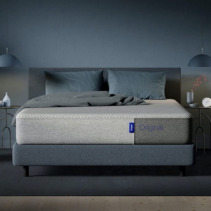 A neatly made bed with gray sheets and pillows rests between two side tables in a dimly lit bedroom. Text: 