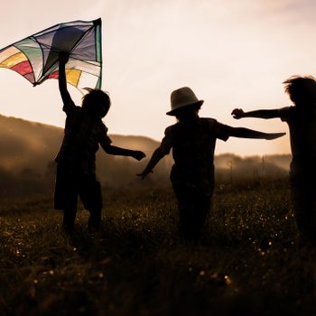 Children run joyfully with a kite in hand, silhouetted against a sunset on a grassy hill.