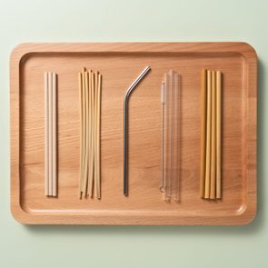 Various reusable straws arranged neatly on a wooden tray against a pale green background.