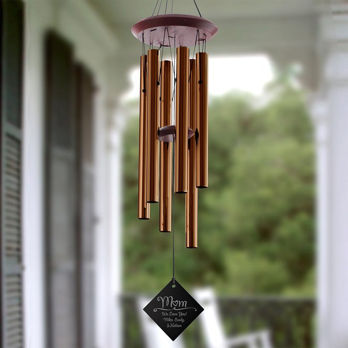 Wind chime hangs, gently swaying on porch, surrounded by blurred greenery. Text on label: 