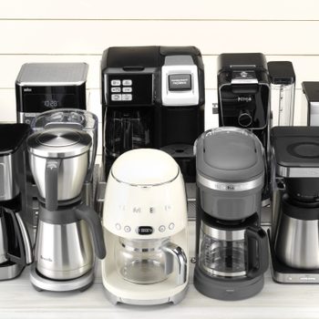 Various coffee makers are displayed on a white wooden surface, showcasing different designs and brands, including Smeg and KitchenAid.