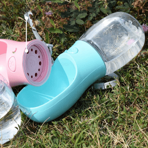 A blue portable pet water bottle lies on grassy ground, next to a pink one, surrounded by greenery and dried plants.