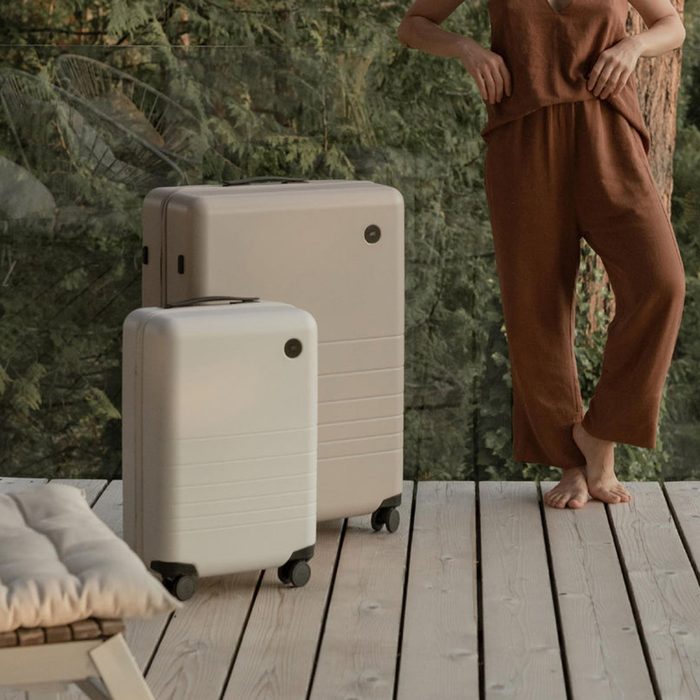 Two beige suitcases stand upright on a wooden deck, with a person in brown clothing nearby, surrounded by greenery.