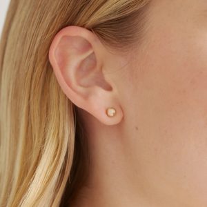 Earring decorates earlobe, featuring a small, round, golden bead; blonde hair frames the ear gently, set against a neutral background.