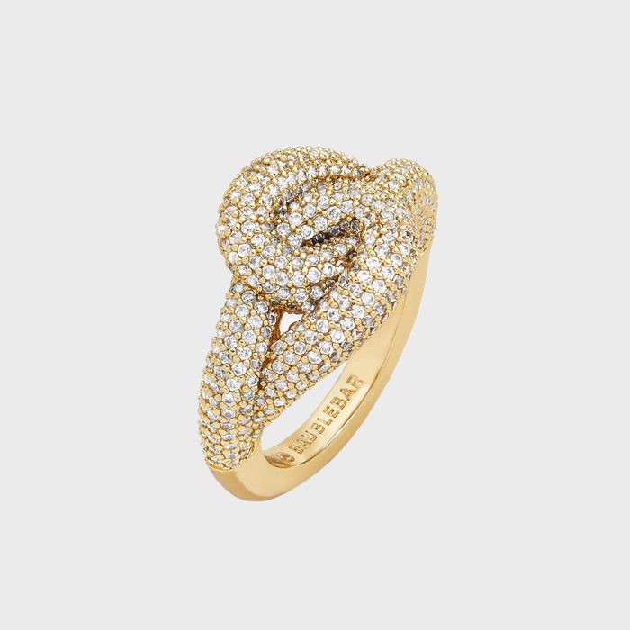 A gold ring encrusted with diamonds forms a knot design. The smooth backdrop highlights the intricate detailing.