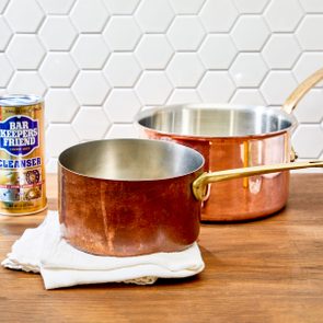 Copper pots rest on a wooden countertop, surrounded by cleaning products. A hexagonal tiled backsplash serves as the background.