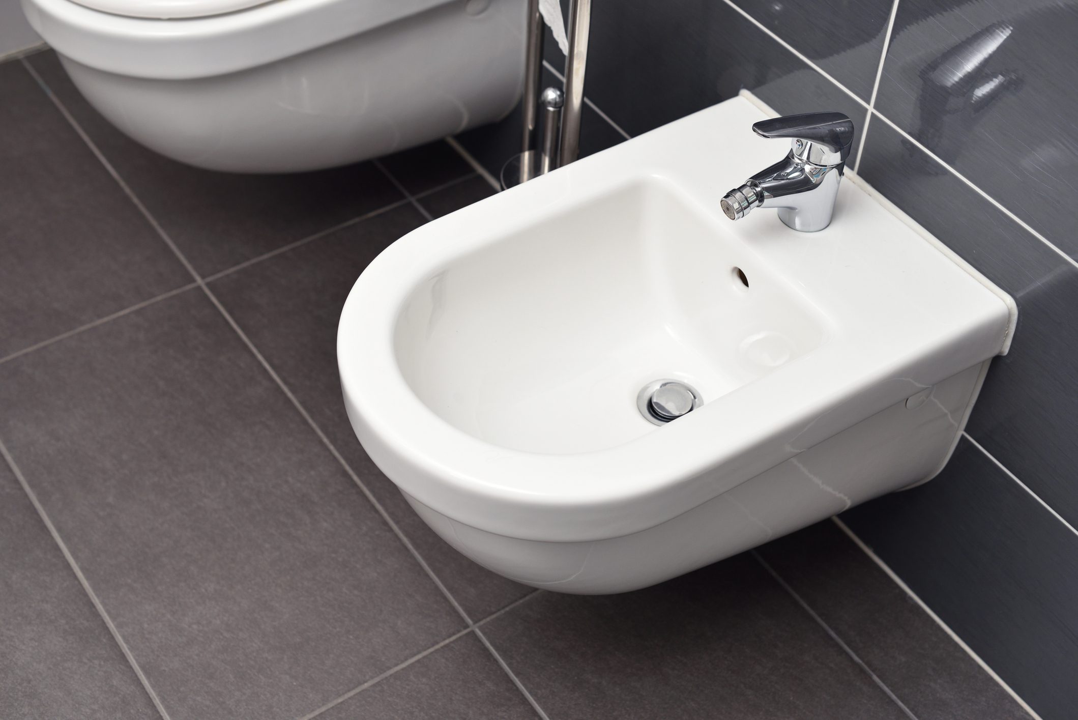 White bidet screwed to a gray tiled wall in the bathroom, close-up