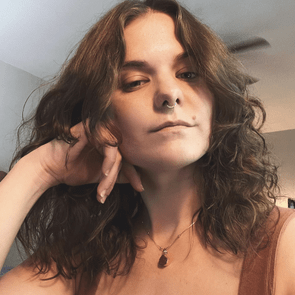 Person resting chin on hand, gazing directly, with curly hair, wearing a necklace, in an indoor, softly lit room with neutral tones.