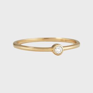 Gold ring displays a small, centered diamond set in a minimalist bezel, against a plain white background.