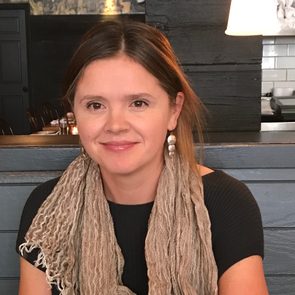 A woman, smiling, sits at a restaurant table, wearing a beige scarf and pearl earrings, with dim lighting and wooden decor surrounding her.