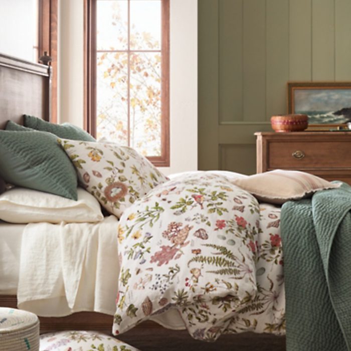 Bed covered with floral-patterned bedding set, adorned with green and beige pillows, situated near a window and wooden dresser in a cozy bedroom.