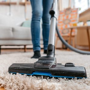 Vacuum cleaner suctioning carpet in a living room, with blurred furniture and person wearing jeans in the background.