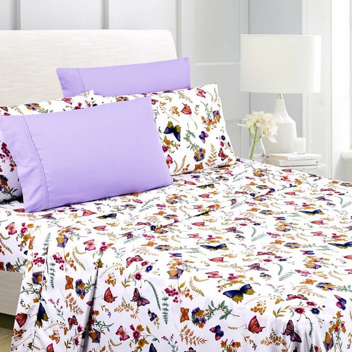 Bedding displays a floral and butterfly pattern, accompanied by solid lavender pillows, on a bed in a bright room with a lamp and flowers.