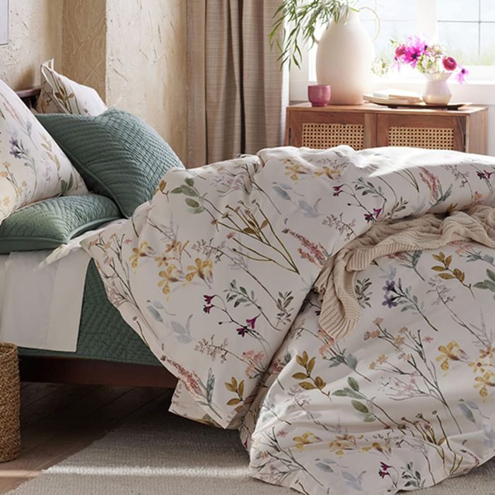 Floral bedding covers a bed, surrounded by green pillows and a wooden nightstand with a vase, books, and flowers in a sunlit room.