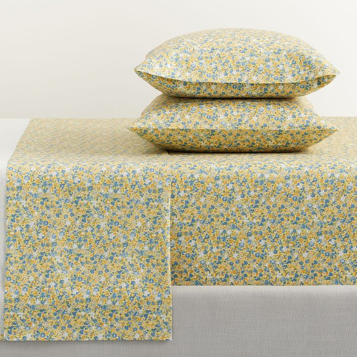 Floral-patterned bedsheets and two pillows lie stacked in a minimal setting, featuring yellow and blue flowers against a muted room background.