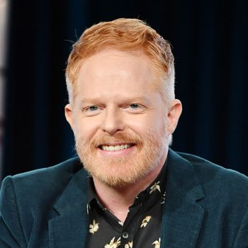 Smiling person with red hair and beard wearing a floral shirt and blue jacket, seated against a blurred dark background.