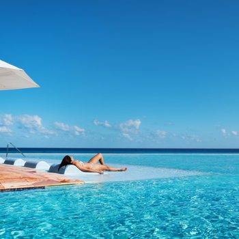 Person sunbathes on pool lounger, surrounded by water and white umbrellas, set against a backdrop of clear blue sky and ocean horizon.