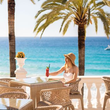 A woman sips a cocktail on a beachside terrace, surrounded by wicker chairs, palm trees, and a distant view of the ocean with a boat.