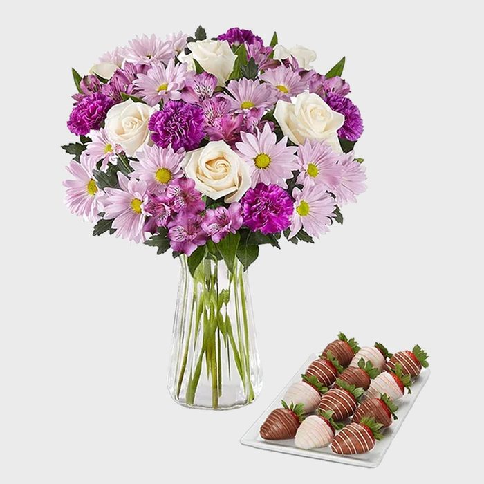 Bouquet of roses and daisies in a clear vase beside a tray of chocolate-covered strawberries in a simple setting.