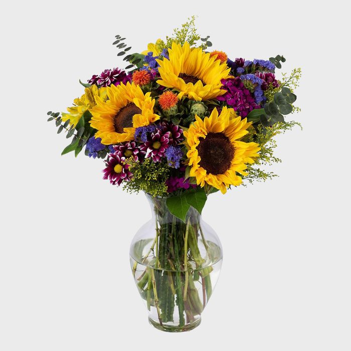 A bouquet of sunflowers and mixed flowers arranged in a clear glass vase, set against a plain white background.