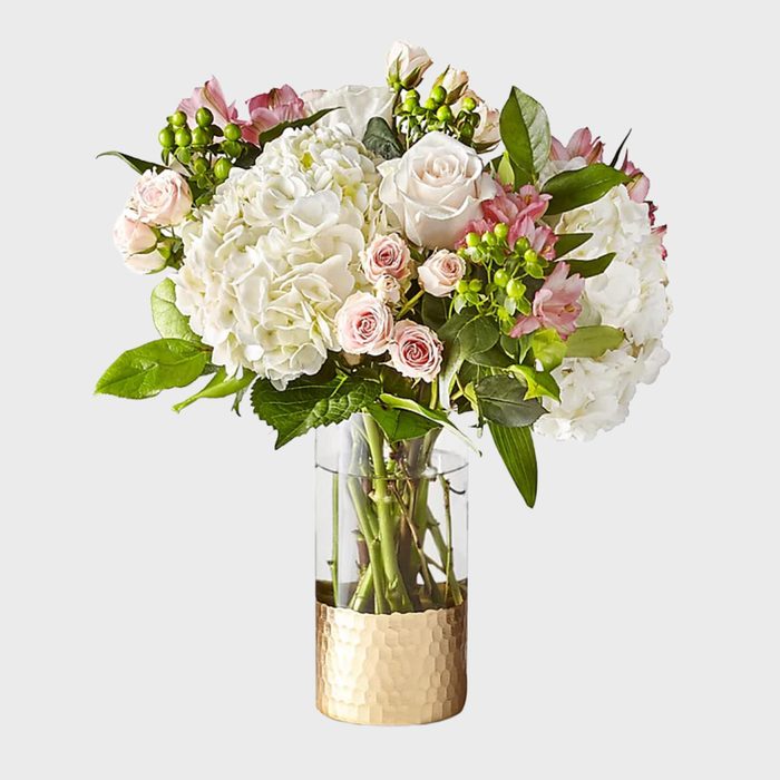 A bouquet of white and pink flowers stands elegantly in a clear vase with a golden base, against a plain background.