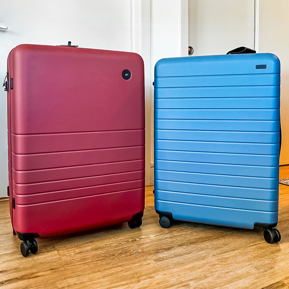 Two upright wheeled suitcases stand on a wooden floor; one is red with a small logo, and the other is blue with horizontal ridges.